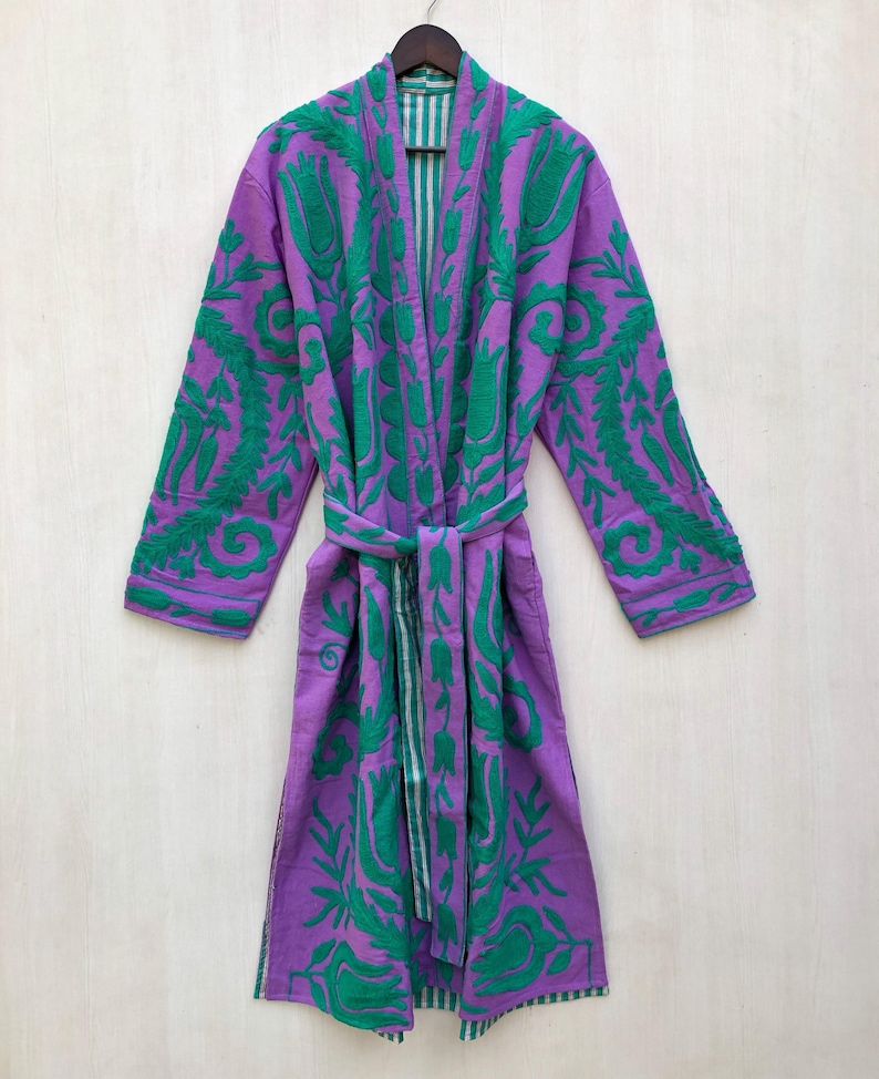 May include: A purple robe with green floral embroidery and a matching belt hangs on a wooden hanger. The robe's interior features a striped pattern. The embroidery covers the sleeves, lapels, and front panels, creating a striking visual contrast.