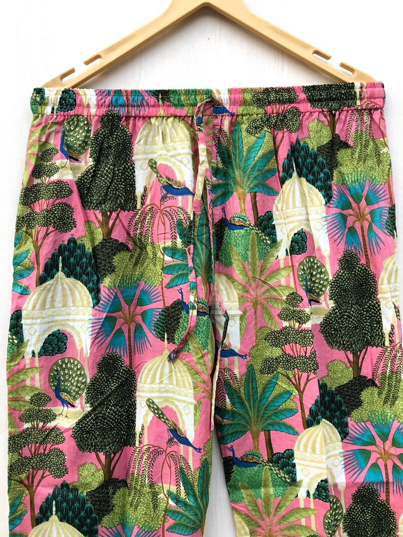 Pu&ograve; includere: Pantaloni da pigiama rosa con una stampa colorata di pavoni, alberi ed edifici bianchi. I pantaloni hanno una cintura elastica e un cordoncino.