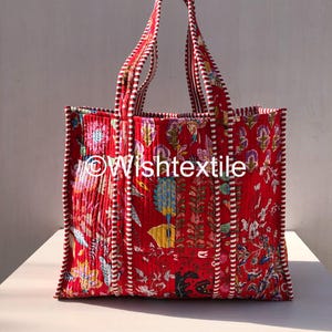 May include: A quilted tote bag in a vibrant red fabric featuring a multi-coloured floral and botanical print. The bag has two handles and vertical piping with a white and red striped pattern. The text Wishtextile is printed on the front.