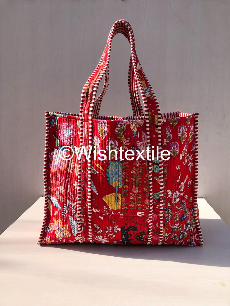 May include: A quilted tote bag in a vibrant red fabric featuring a multi-coloured floral and botanical print. The bag has two handles and vertical piping with a white and red striped pattern. The text Wishtextile is printed on the front.