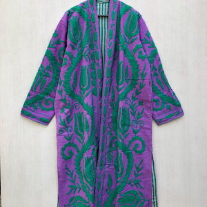 May include: A long, purple robe with green floral embroidery. The robe features a striped lining and is displayed on a wooden hanger. The embroidery design includes stylized flowers and leaves, creating a decorative pattern across the garment.