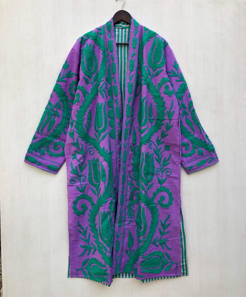 May include: A long, purple robe with green floral embroidery. The robe features a striped lining and is displayed on a wooden hanger. The embroidery design includes stylized flowers and leaves, creating a decorative pattern across the garment.
