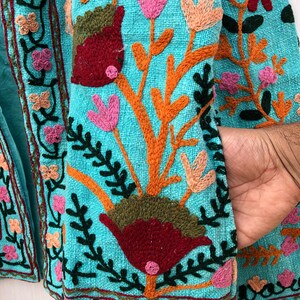 May include: A turquoise fabric jacket with colorful floral embroidery. The embroidery features pink, red, orange, green, and brown thread. The jacket has a pocket on the right side.