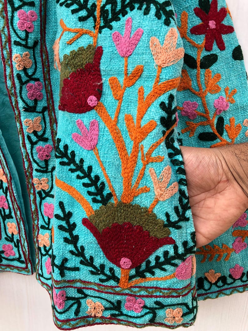 May include: A turquoise fabric jacket with colorful floral embroidery. The embroidery features pink, red, orange, green, and brown thread. The jacket has a pocket on the right side.