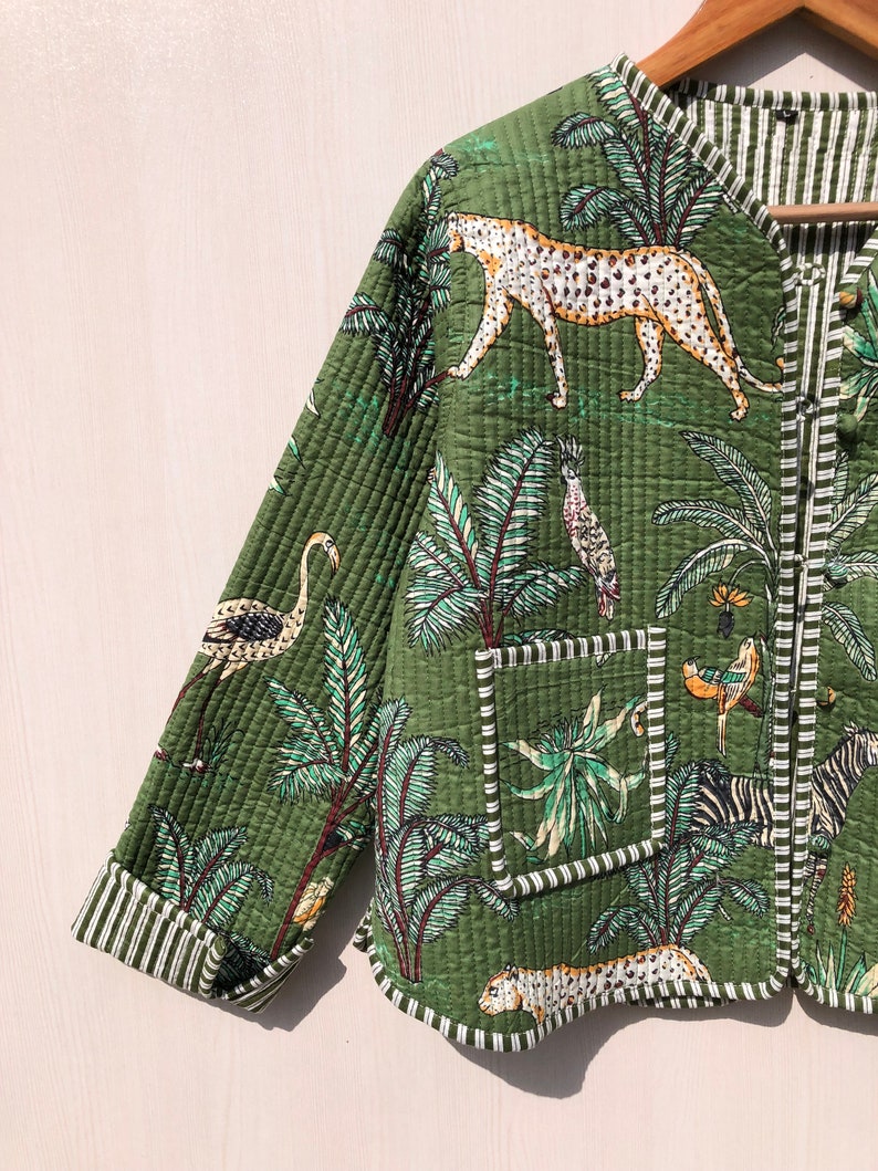 May include: A green quilted jacket with a jungle print featuring leopards, zebras, birds, and palm trees. The jacket has a white and green striped lining and a white and green striped pocket.