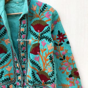 May include: A turquoise blue jacket with floral embroidery in shades of pink, orange, green, and red. The jacket has a collar and a button closure.
