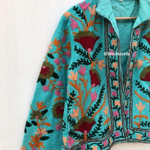 May include: A turquoise blue jacket with a floral embroidery design. The jacket has a collar and a front closure. The embroidery is in shades of pink, red, orange, green, and brown. The text "@Wishtextile" is visible on the jacket.