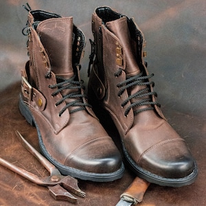 May include: A pair of brown leather boots with black laces and buckles. The boots are on a brown leather surface with a pair of pliers and a knife.