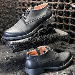 May include: Two pairs of black leather lace-up shoes with a cap toe design. The shoes are on a dark surface.