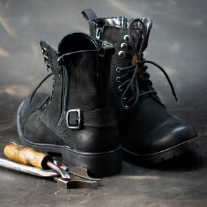 K&ouml;nnte beinhalten: Zwei schwarze Lederstiefel mit Schnallen und Schnallen, die auf einer dunkelbraunen Oberfl&auml;che mit einem Metallwerkzeug liegen.