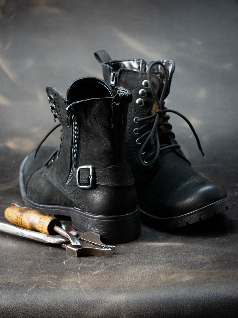 K&ouml;nnte beinhalten: Zwei schwarze Lederstiefel mit Schnallen und Schnallen, die auf einer dunkelbraunen Oberfl&auml;che mit einem Metallwerkzeug liegen.