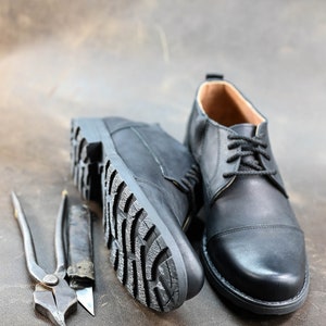 May include: A pair of black leather shoes with laces. One shoe is shown with the sole facing the camera, while the other shoe is shown from the side. The shoes are on a brown leather surface.