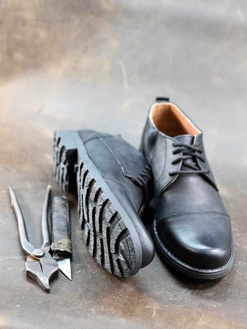 May include: A pair of black leather shoes with laces. One shoe is shown with the sole facing the camera, while the other shoe is shown from the side. The shoes are on a brown leather surface.