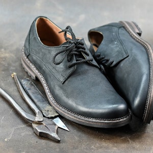 May include: Two black leather shoes with laces, one with the laces tied, and a pair of metal pliers on a brown surface.