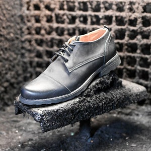 May include: A black leather oxford shoe with a lace-up closure. The shoe is sitting on a rough, black surface.