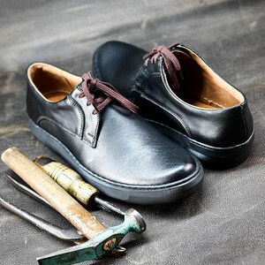 May include: Two black leather shoes with brown laces and a white sole. The shoes are on a gray surface with a hammer and other tools.