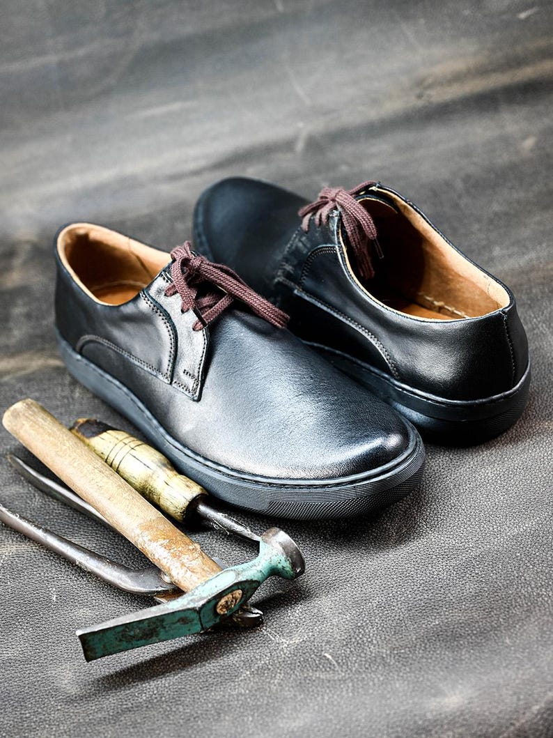 May include: Two black leather shoes with brown laces and a white sole. The shoes are on a gray surface with a hammer and other tools.