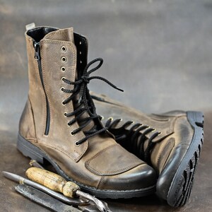 May include: A pair of brown leather boots with black laces and a zipper on the side. The boots are sitting on a brown surface with two leather working tools.