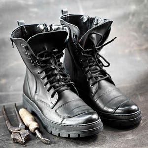 Puede incluir: Un par de botas de cuero negro con cordones y una cremallera en el lateral. Las botas tienen una suela gruesa y est&aacute;n sobre una superficie gris.