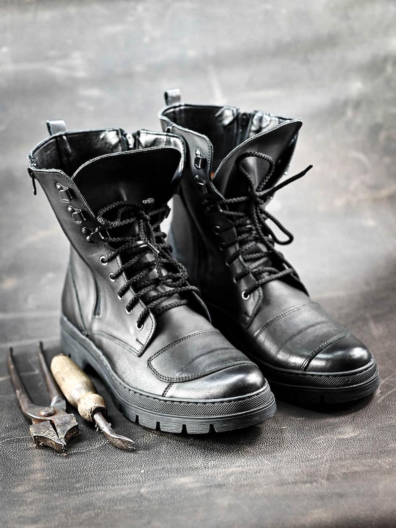 Puede incluir: Un par de botas de cuero negro con cordones y una cremallera en el lateral. Las botas tienen una suela gruesa y est&aacute;n sobre una superficie gris.