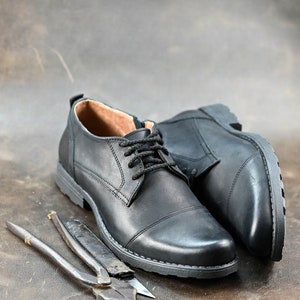 May include: Two black leather shoes with laces and a pair of pliers on a brown leather surface.