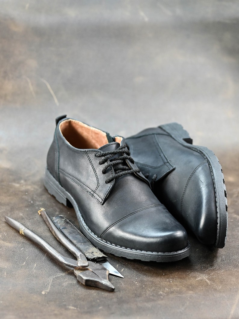 May include: Two black leather shoes with laces and a pair of pliers on a brown leather surface.
