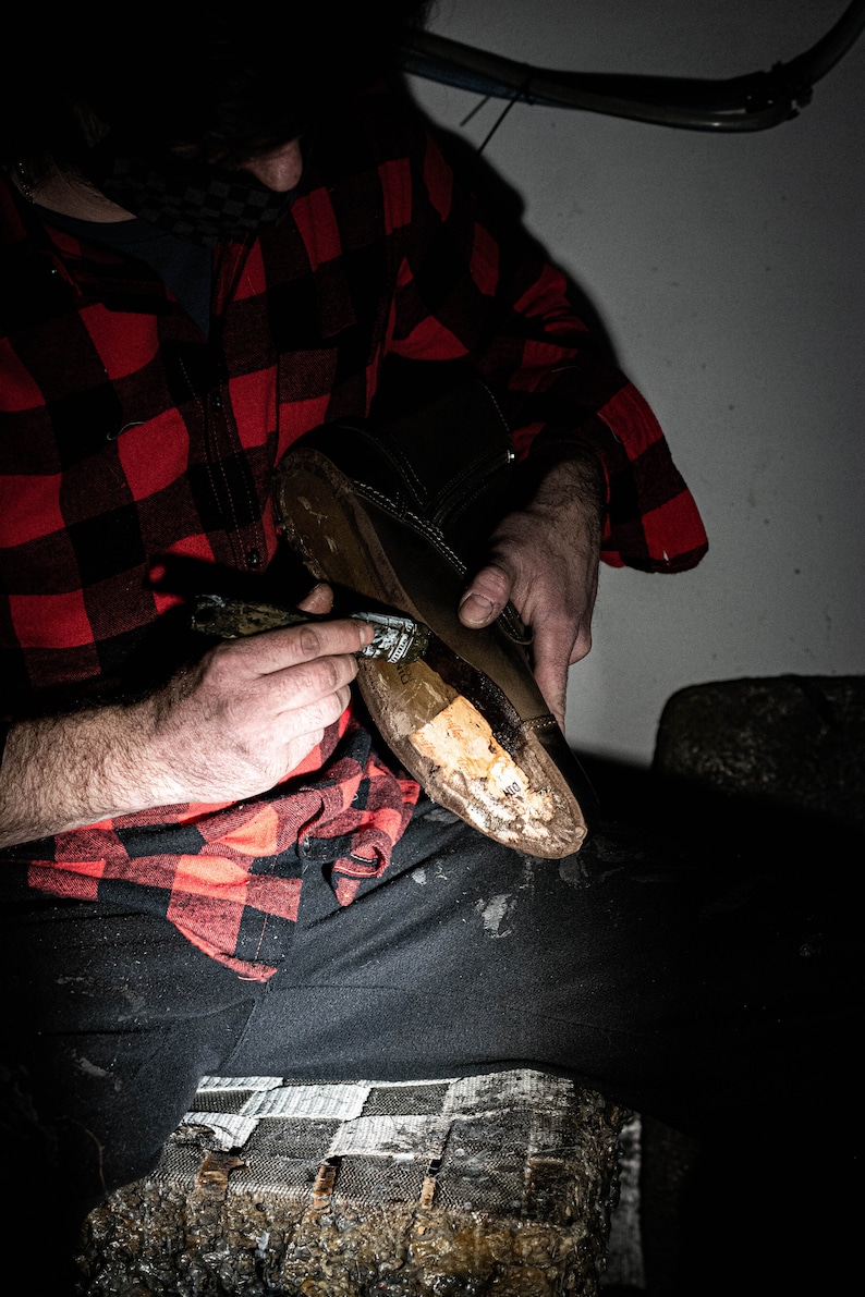 May include: A person wearing a red and black plaid shirt is working on a brown leather boot. They are using a tool to apply a substance to the boot.