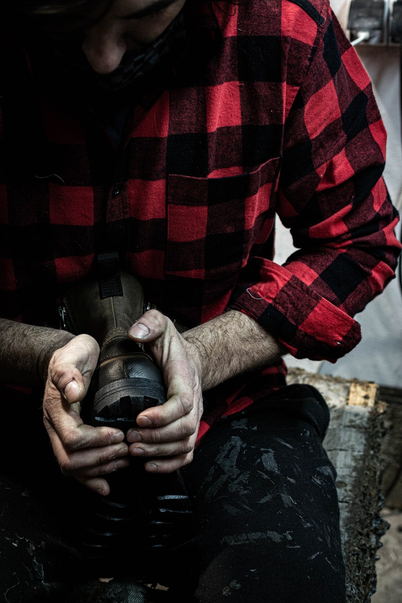May include: A person wearing a red and black plaid shirt is holding a brown leather boot in their hands. The boot has a black sole and laces.