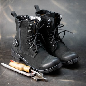 K&ouml;nnte beinhalten: Ein Paar schwarze Lederstiefel mit Schn&uuml;rsenkeln und einem Rei&szlig;verschluss an der Seite. Die Stiefel stehen auf einer braunen Oberfl&auml;che mit einem Lederbearbeitungswerkzeug.