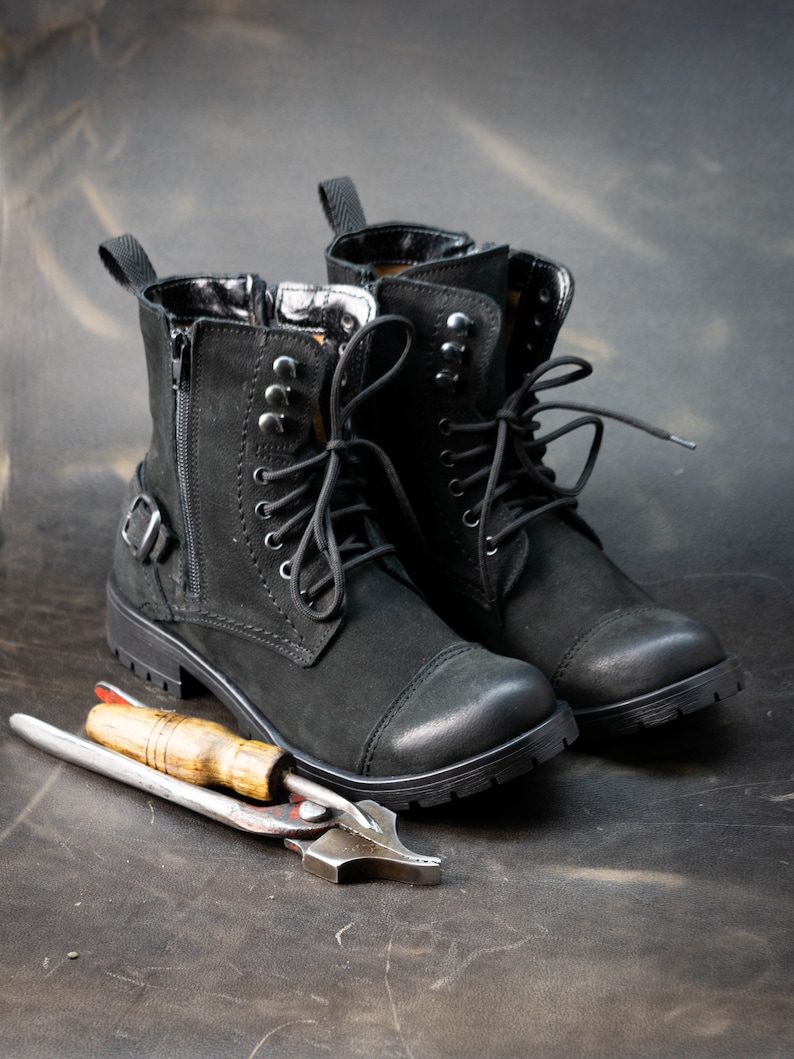 K&ouml;nnte beinhalten: Ein Paar schwarze Lederstiefel mit Schn&uuml;rsenkeln und einem Rei&szlig;verschluss an der Seite. Die Stiefel stehen auf einer braunen Oberfl&auml;che mit einem Lederbearbeitungswerkzeug.