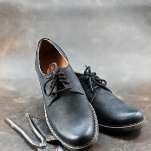 May include: Two black leather lace-up shoes with a brown leather background. The shoes are sitting next to two metal tools.