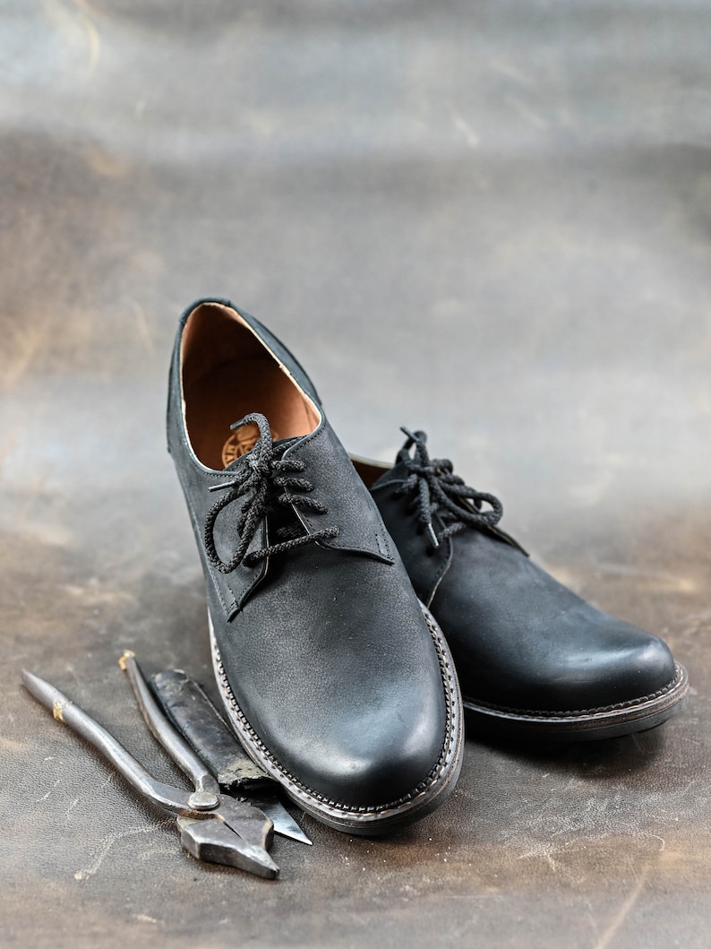 May include: Two black leather lace-up shoes with a brown leather background. The shoes are sitting next to two metal tools.