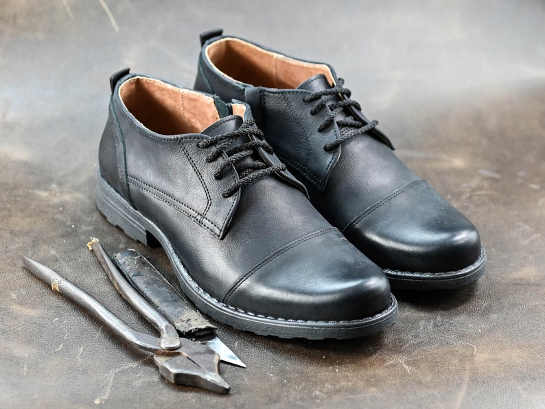 May include: Two black leather lace-up shoes with a cap toe design. The shoes are on a brown surface with two metal tools.