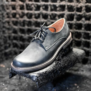 May include: A black leather oxford shoe with a lace-up closure. The shoe is sitting on a black, textured surface.