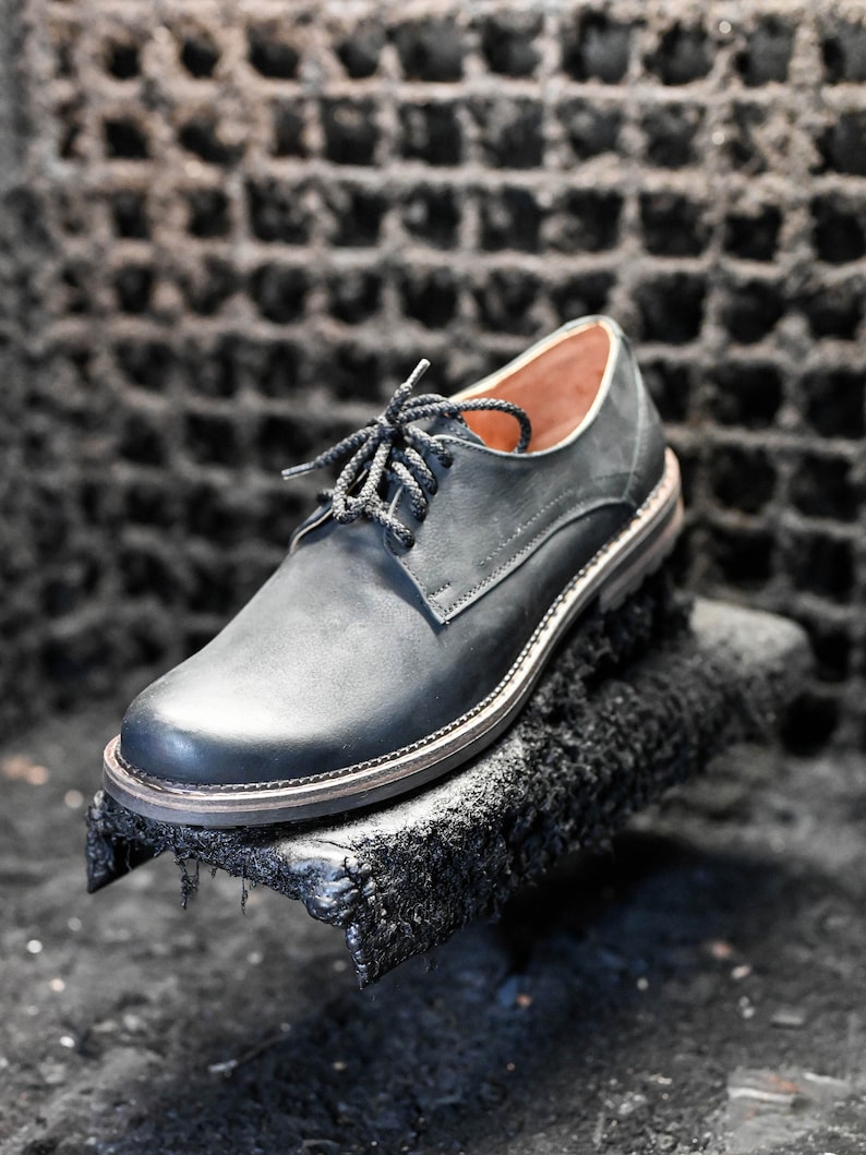 May include: A black leather oxford shoe with a lace-up closure. The shoe is sitting on a black, textured surface.