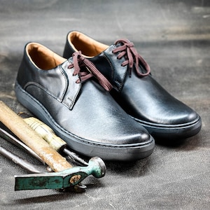 May include: Two black leather lace-up shoes with brown laces on a gray textured surface. The shoes have a white rubber sole. There are three tools, including a hammer, lying in front of the shoes.