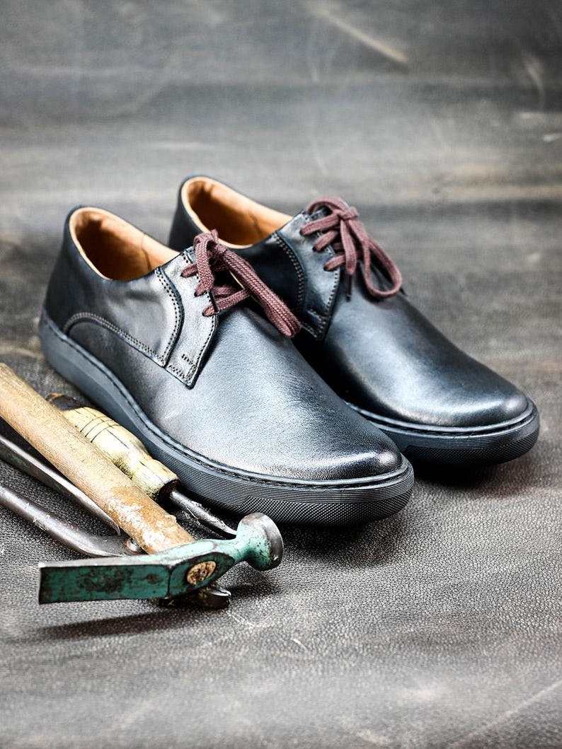 May include: Two black leather lace-up shoes with brown laces on a gray textured surface. The shoes have a white rubber sole. There are three tools, including a hammer, lying in front of the shoes.