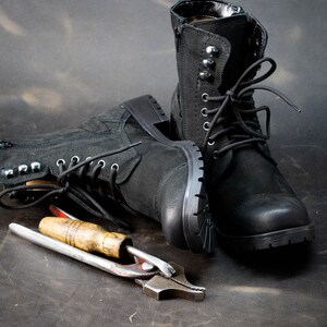 K&ouml;nnte beinhalten: Zwei schwarze Lederstiefel mit Schn&uuml;rsenkeln und Metallverzierungen, die auf einer braunen Oberfl&auml;che mit einem Werkzeug aus Metall mit Holzgriff liegen.