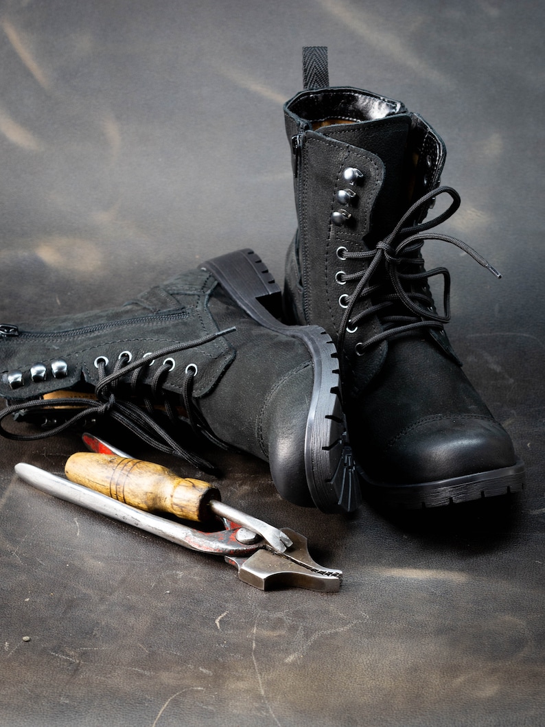 K&ouml;nnte beinhalten: Zwei schwarze Lederstiefel mit Schn&uuml;rsenkeln und Metallverzierungen, die auf einer braunen Oberfl&auml;che mit einem Werkzeug aus Metall mit Holzgriff liegen.
