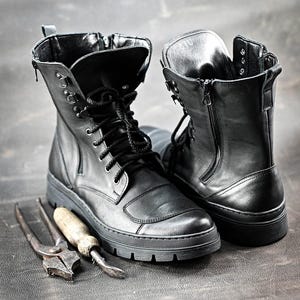 Puede incluir: Dos botas de cuero negro con cordones y cremalleras. Las botas est&aacute;n sobre una superficie gris con dos herramientas de metal.
