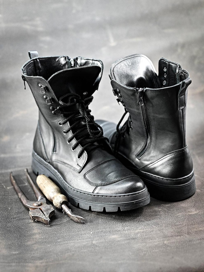 Puede incluir: Dos botas de cuero negro con cordones y cremalleras. Las botas est&aacute;n sobre una superficie gris con dos herramientas de metal.