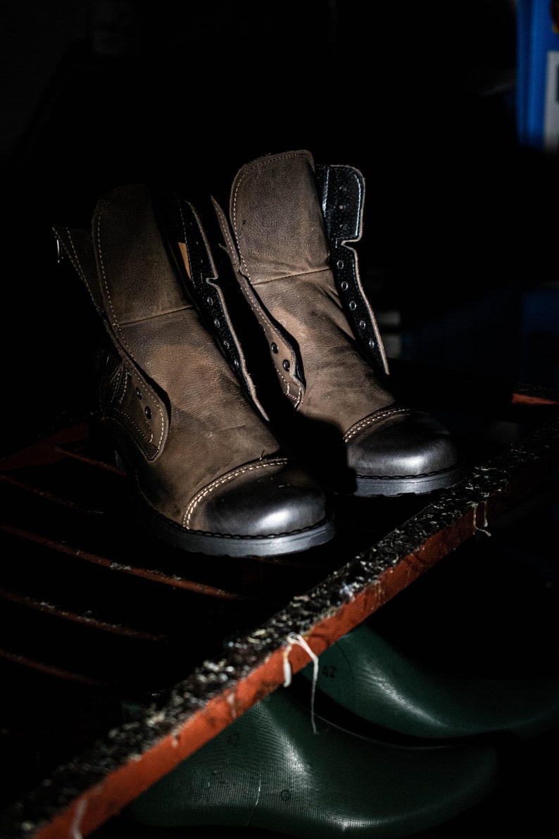 May include: Two brown leather boots with black laces and black soles. The boots are sitting on a wooden shelf.