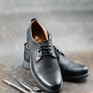 May include: A pair of black leather lace-up shoes with a cap toe design. The shoes are on a brown surface with two metal tools.