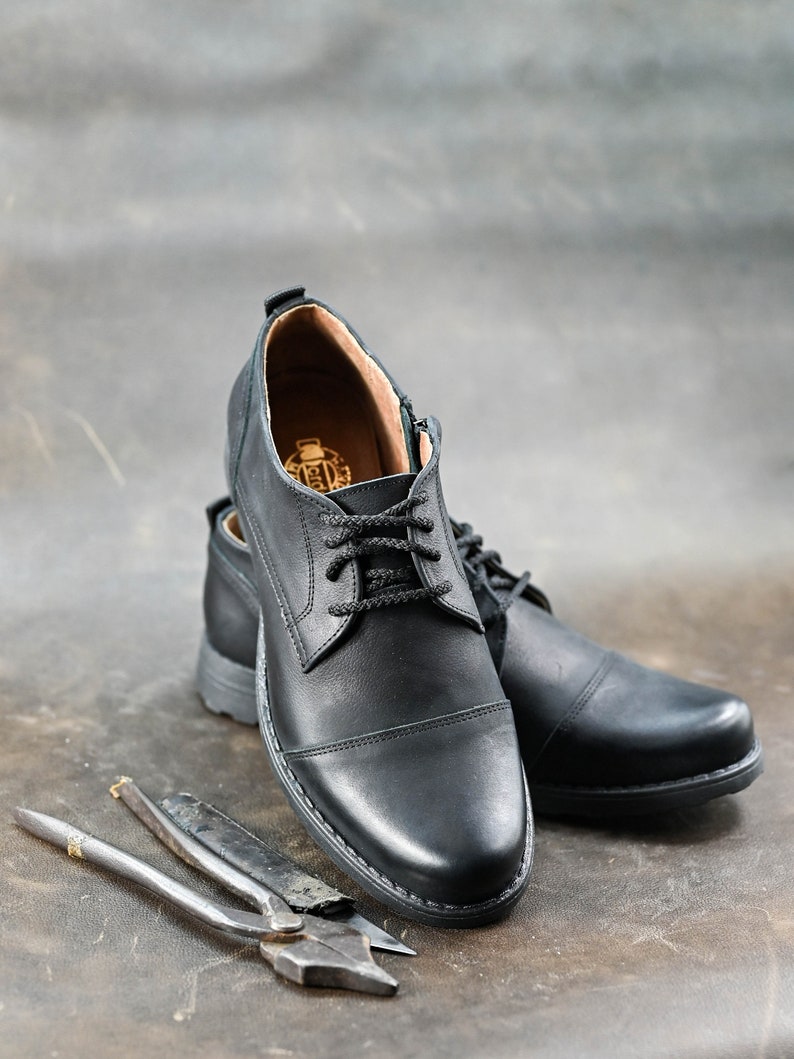 May include: A pair of black leather lace-up shoes with a cap toe design. The shoes are on a brown surface with two metal tools.