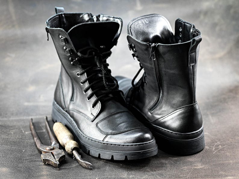 Puede incluir: Dos botas de cuero negro con cordones y cremalleras. Una bota tiene una cremallera en el lateral y la otra tiene cordones. Las botas est&aacute;n sobre una superficie gris texturizada.