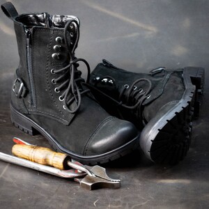 K&ouml;nnte beinhalten: Ein Paar schwarze Lederstiefel mit Schn&uuml;rsenkeln und Schnallen. Die Stiefel befinden sich auf einer braunen Oberfl&auml;che mit einem Metallwerkzeug.