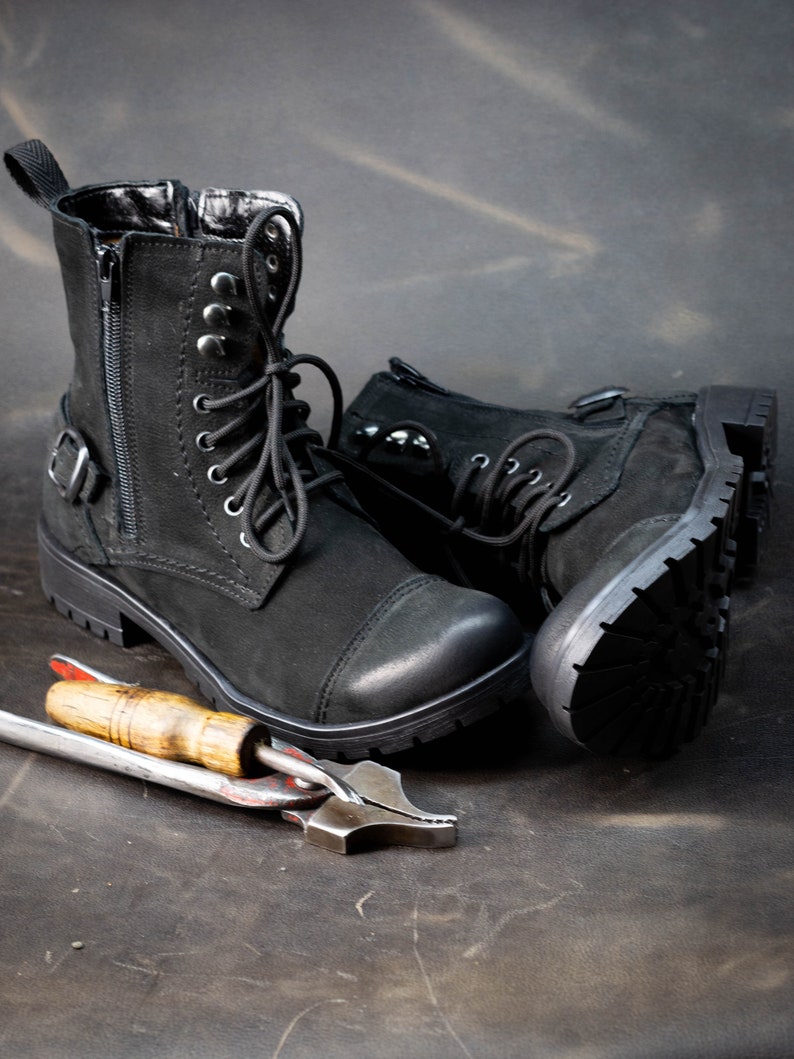 K&ouml;nnte beinhalten: Ein Paar schwarze Lederstiefel mit Schn&uuml;rsenkeln und Schnallen. Die Stiefel befinden sich auf einer braunen Oberfl&auml;che mit einem Metallwerkzeug.