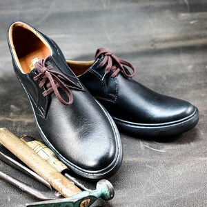 May include: Two black leather lace-up shoes with brown laces. The shoes are sitting on a gray textured surface with a hammer and other tools.