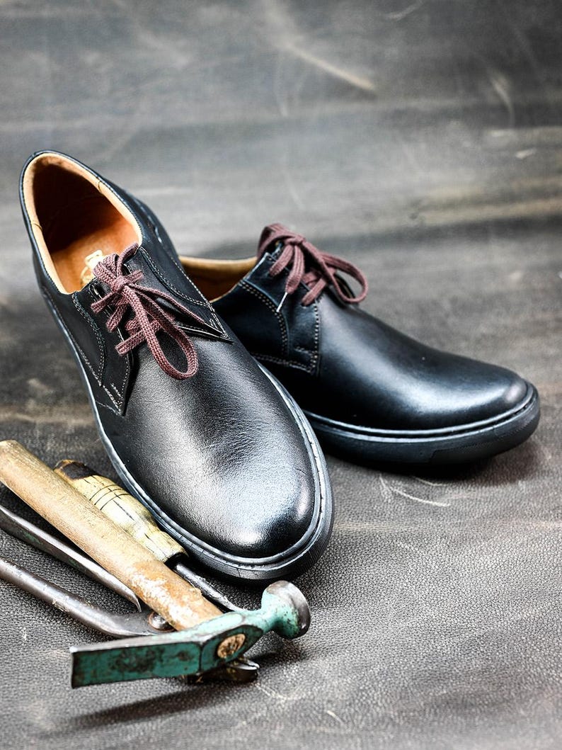 May include: Two black leather lace-up shoes with brown laces. The shoes are sitting on a gray textured surface with a hammer and other tools.
