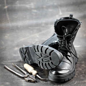 Puede incluir: Botas de cuero negro con cordones y una suela gruesa de goma. Las botas est&aacute;n sobre una superficie gris oscuro con un par de alicates y un destornillador.